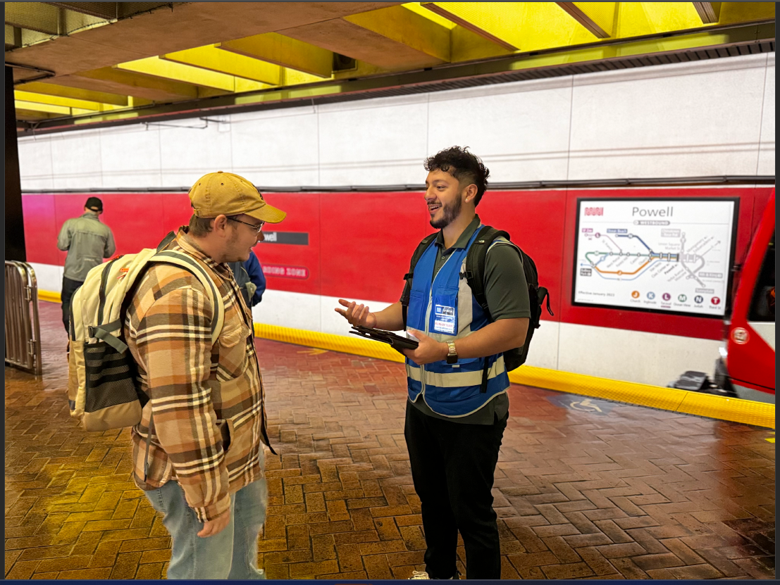 Survey taker wearing a blue vest and badge speaks with a Muni rider on the Powell Station platform for the onboard ridership survey.